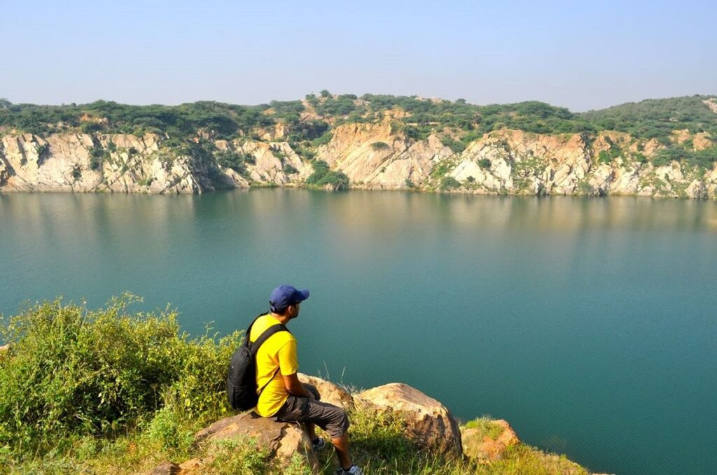 A man wearing a yellow shirt and blue cap sits on a rock, overlooking the serene blue waters of Bhardwaj Lake, surrounded by rocky cliffs and greenery in Delhi’s Asola Bhatti Wildlife Sanctuary.