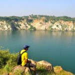 A man wearing a yellow shirt and blue cap sits on a rock, overlooking the serene blue waters of Bhardwaj Lake, surrounded by rocky cliffs and greenery in Delhi’s Asola Bhatti Wildlife Sanctuary.