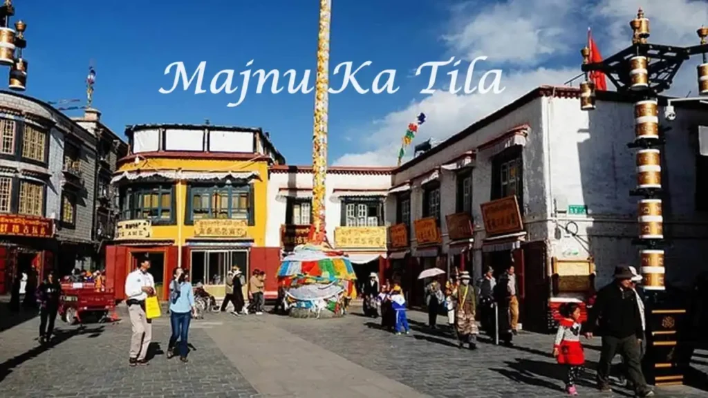 Street view of Majnu Ka Tilla in Delhi, showcasing colorful Tibetan-style buildings, prayer flags, and people walking in the marketplace under a bright blue sky.