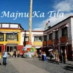 Street view of Majnu Ka Tilla in Delhi, showcasing colorful Tibetan-style buildings, prayer flags, and people walking in the marketplace under a bright blue sky.