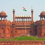 Front view of the historic Red Fort in Old Delhi, India, with the Indian national flag flying atop its central dome and red sandstone walls under a clear sky.