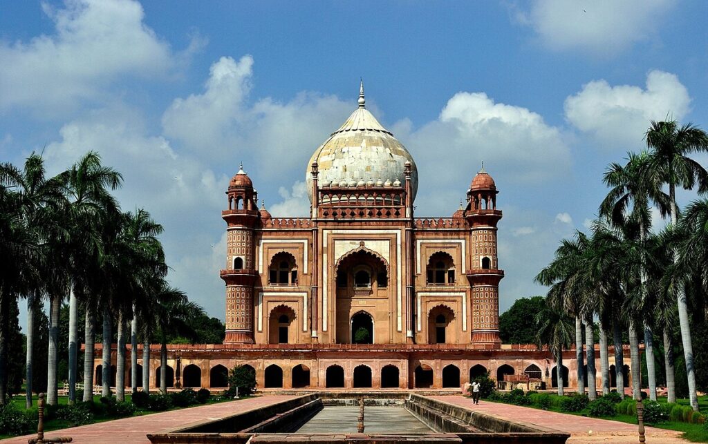 Front view of Safdarjung Tomb in Delhi, featuring Mughal-style red sandstone architecture, a large central dome, palm trees on both sides, and a water channel leading to the monument under a partly cloudy blue sky.