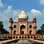 Front view of Safdarjung Tomb in Delhi, featuring Mughal-style red sandstone architecture, a large central dome, palm trees on both sides, and a water channel leading to the monument under a partly cloudy blue sky.