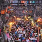 A bustling street scene in Chandni Chowk, Delhi, filled with vibrant shops, tangled overhead wires, and a dense crowd of people exploring the market.