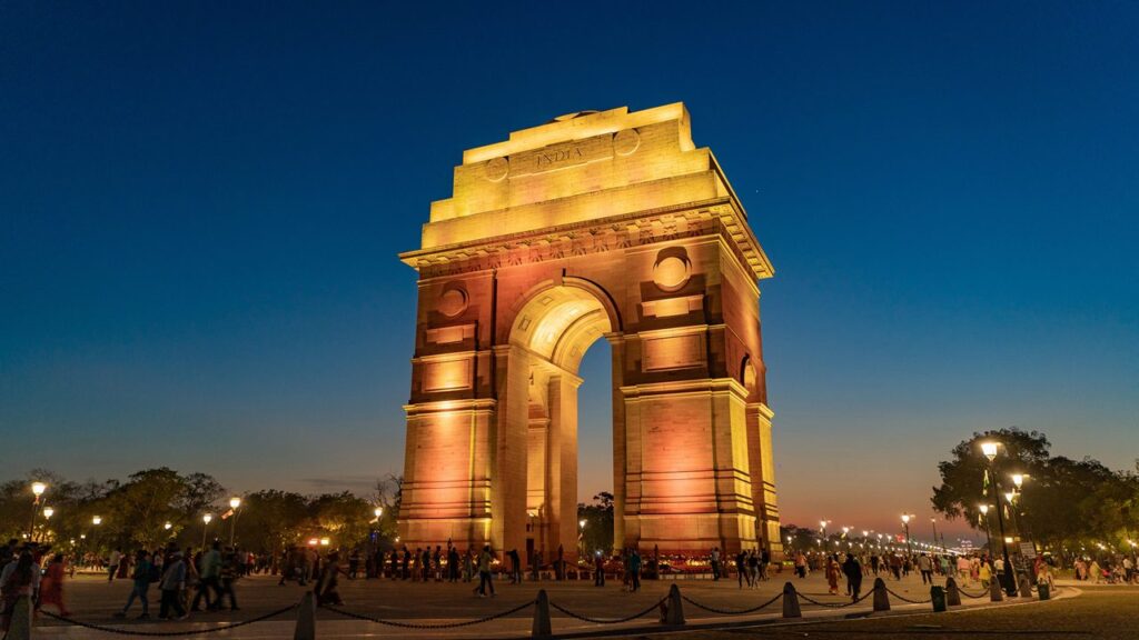 India Gate in Delhi illuminated at night with visitors walking around the monument against a twilight sky.