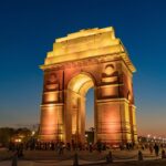 India Gate in Delhi illuminated at night with visitors walking around the monument against a twilight sky.