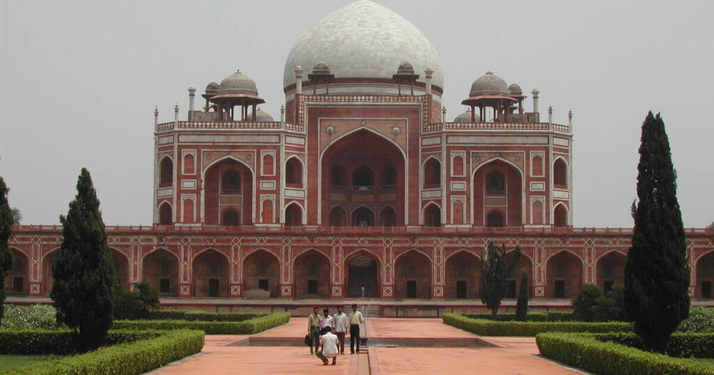 Front view of Humayun’s Tomb in Delhi showcasing its grand Mughal architecture with red sandstone arches, white marble dome, and lush green gardens.